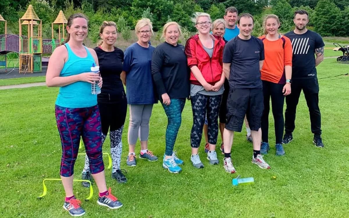 A group of smiling clients and a trainer standing together on the grass in a park after an outdoor session
